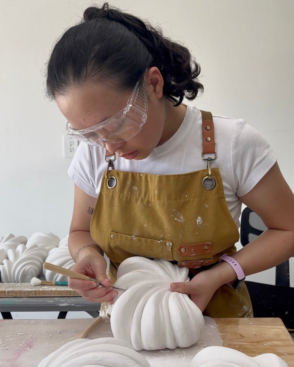 A woman wearing safety glasses and a mustard yellow apron over a white t-shirt, carefully painting white, ribbed decorative artwork on a table in a studio setting.