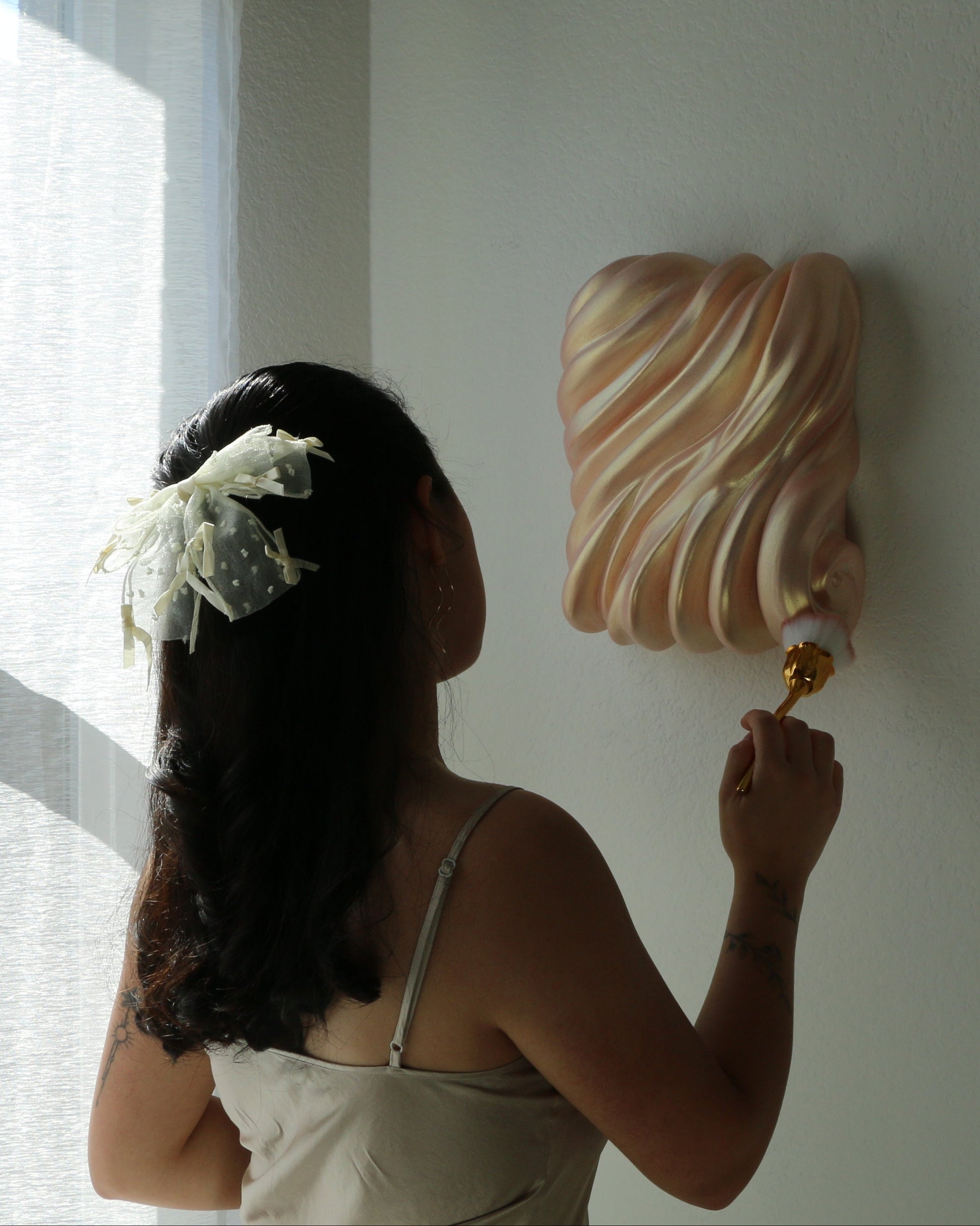 A woman with dark hair and a white flower hair clip is seen from behind, holding a paint roller and applying white paint to a textured wall. She is wearing a white spaghetti-strap top.