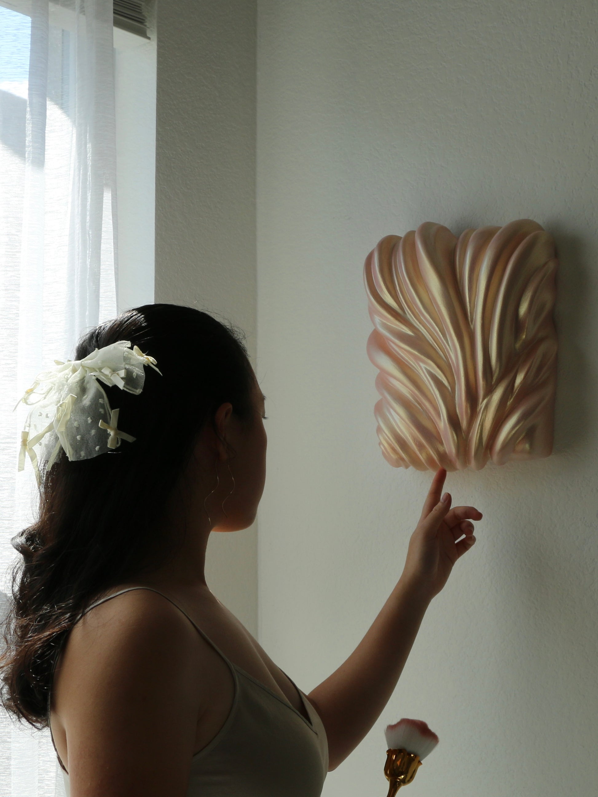 A woman in a beige dress points at a decorative, shell-like wall hanging mounted on a white wall, with soft window light on the left.